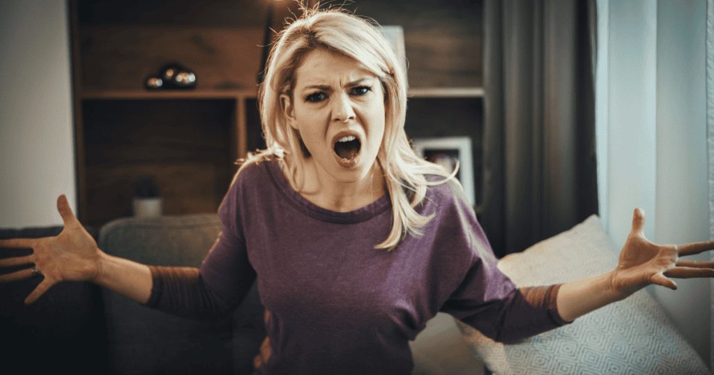 A blonde woman throws her hands up with an open-mouthed, furious shout on a living room couch, showcasing intense, accusatory anger directed into a shared space.