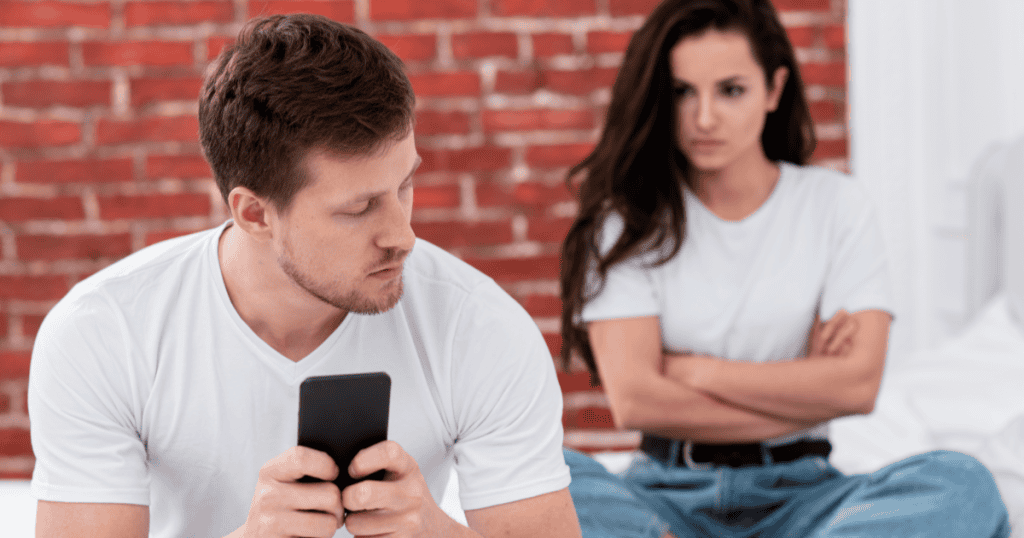 A man focuses on his phone while a woman sits with arms crossed and a distant, disapproving look in the background, illustrating the emotional disconnect behind repeated comparisons to an ex.