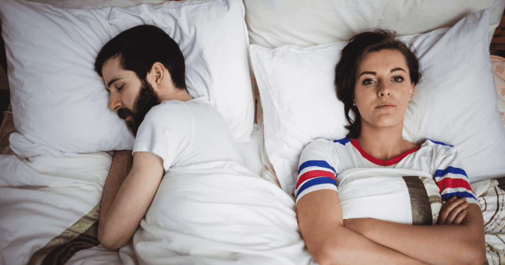 A man sleeping soundly while his partner lies awake with arms crossed and a hollow stare, capturing the emotional isolation a narcissist engineers in the bedroom to maintain control.