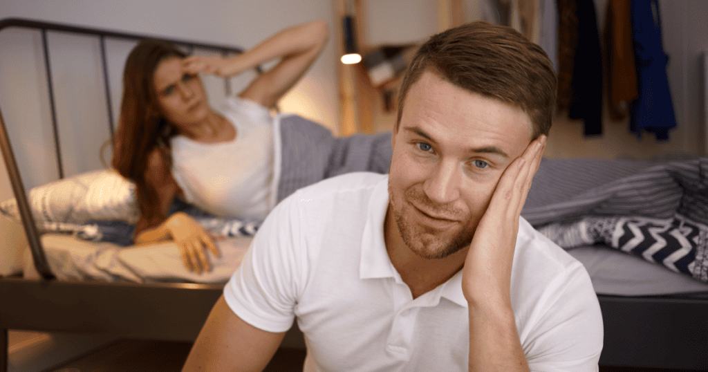 A man sitting smugly detached while his distressed partner lies behind him in bed, illustrating how a narcissist uses emotional withdrawal and indifference in the bedroom as a calculated tool for control.