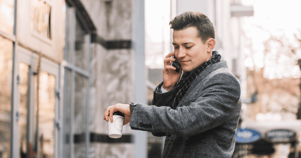 A man on the phone checks his watch while holding a coffee outside, capturing the always-busy excuse used to avoid giving time that feels like too much to give.