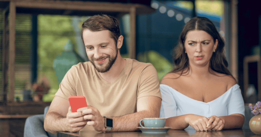 A man smiles at his phone while a woman beside him at the café table looks visibly annoyed, capturing the frustration of being sidelined by go-to excuses.
