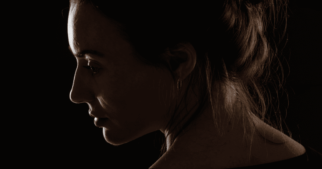 A woman’s face is outlined in dramatic profile against deep darkness with soft rim lighting, capturing the somber introspection and quiet withdrawal that precedes emotional detachment.