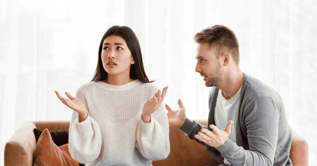 A woman throws up her hands in frustration while a man gestures emphatically during a tense conversation on the couch, reflecting how a comparison pattern began well before the current relationship.