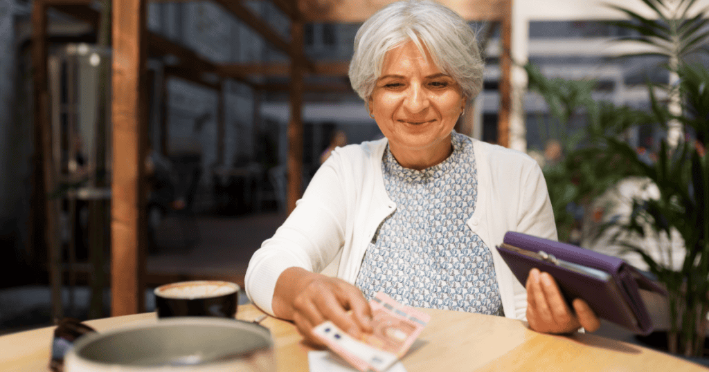 A smiling older woman places cash on a café table while holding her wallet, illustrating how a narcissist's warmth and charm can suddenly switch on the moment money enters the room.