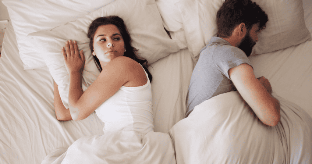 A woman lying awake and unsettled while her partner turns away in bed, depicting how a narcissist's deliberate physical restlessness disrupts their partner's sleep and peace as a subtle form of control.