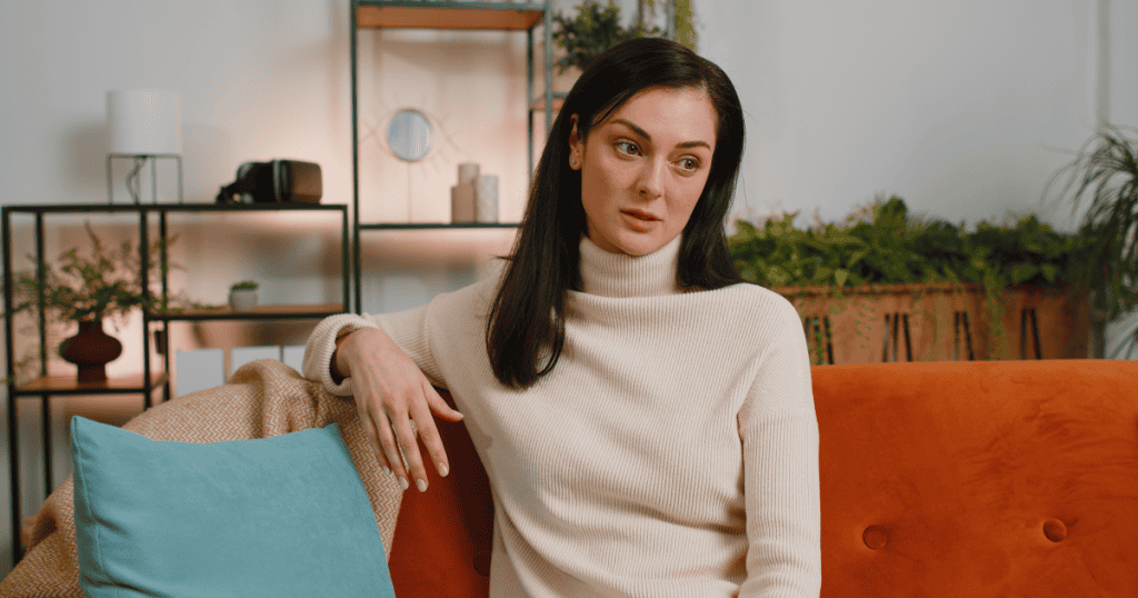 A woman sitting alone on a couch with a hollow, distant stare, capturing the quiet, numb anger of a survivor who has stopped screaming and started simply disconnecting.