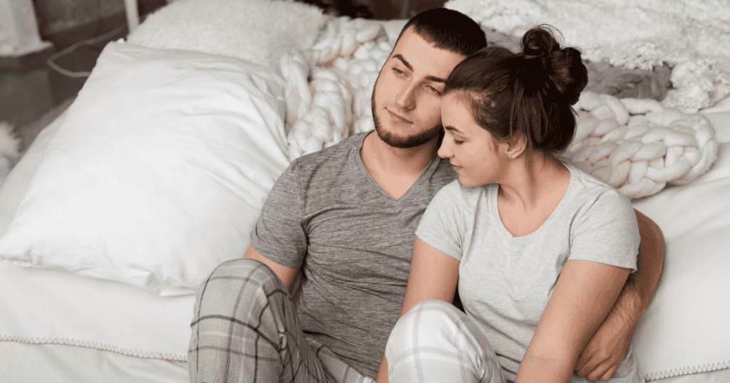 A couple sitting closely in bed with the man appearing calm and calculated, depicting how a narcissist strategically offers physical closeness as a reward to condition their partner into compliance.