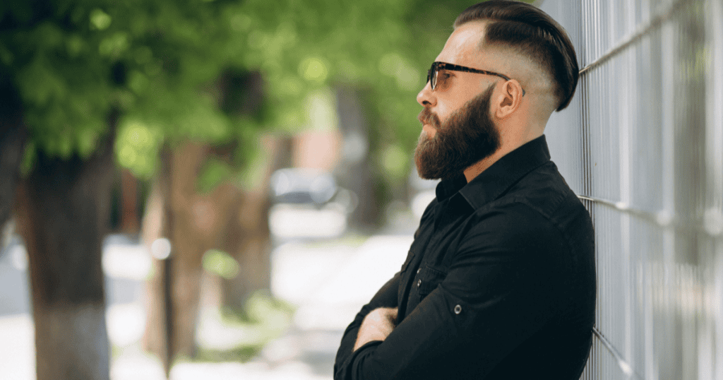 A bearded man in sunglasses leans against a fence with arms crossed, his cool, detached stance reflecting a guarded self-interest devoid of genuine integrity.