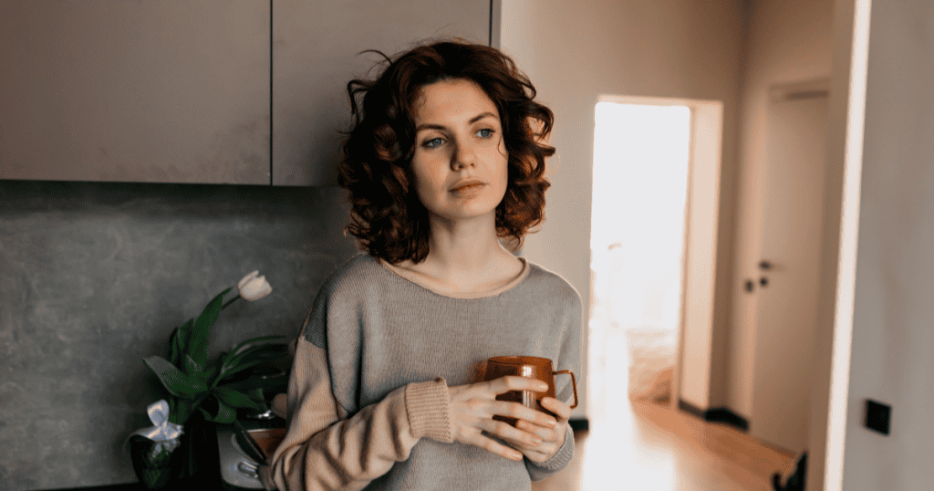 A woman stands alone in her kitchen holding a mug with a pensive, distant expression, reflecting the quiet realization that the comparisons were never a competition but a form of control.
