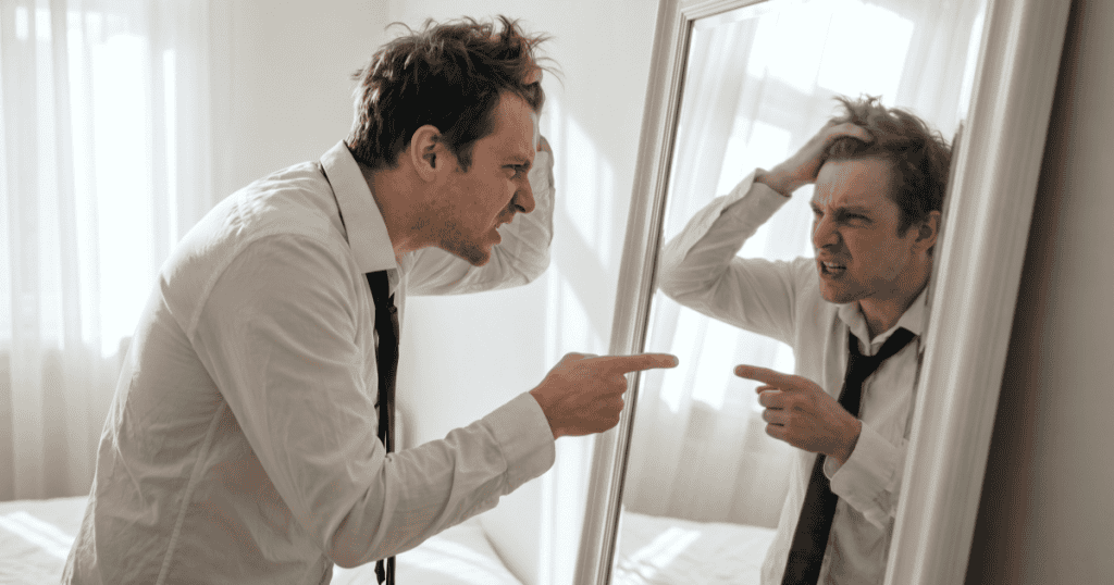 A man grips his hair and points angrily at his own reflection in the mirror, capturing the raw self-hatred that may lie hidden beneath a narcissist's ego.