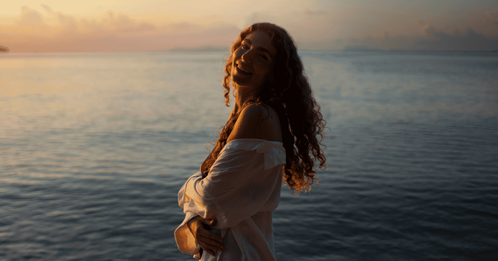 A curly-haired woman smiles warmly over her shoulder while standing in front of a calm ocean at sunset, embodying the radiant peace that comes from stepping entirely outside vulnerable spaces.