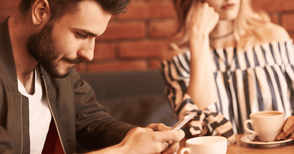 A man looks intently down at his smartphone at a cafe table while a woman rests her chin on her hand in the blurred background, highlighting emotional absence and disengagement in shared spaces.