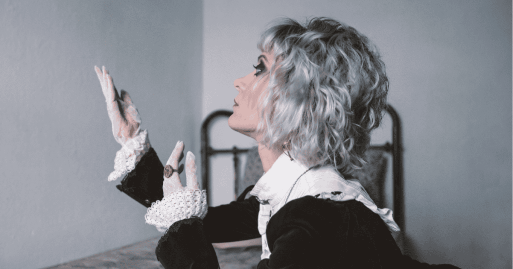 A woman with short silver curls raises lace-gloved hands with dramatic expression in a stark room, portraying the repetitive theatrical performance maintained across every setting.