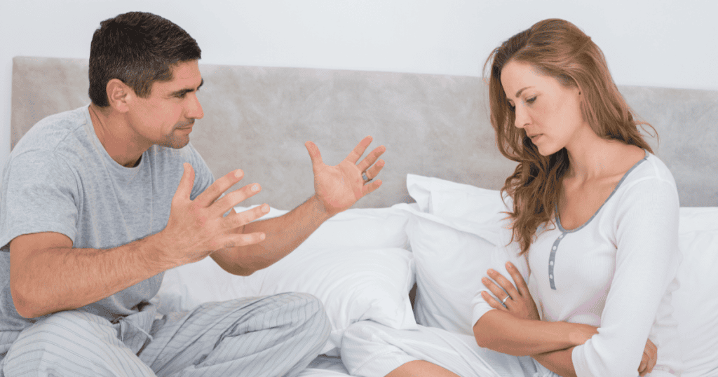 A man gesturing aggressively in bed while his partner sits with arms crossed and eyes down, showing how a narcissist turns the bedroom into a space to exploit your insecurities and keep you emotionally defenseless.