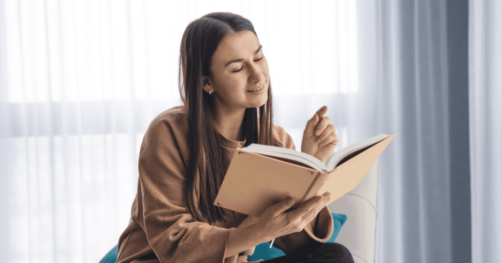 A woman reads aloud from an open hardcover book with an expressive, knowing gesture in front of sheer curtains, illustrating the clarity and empowerment found in understanding the language of covert dynamics.