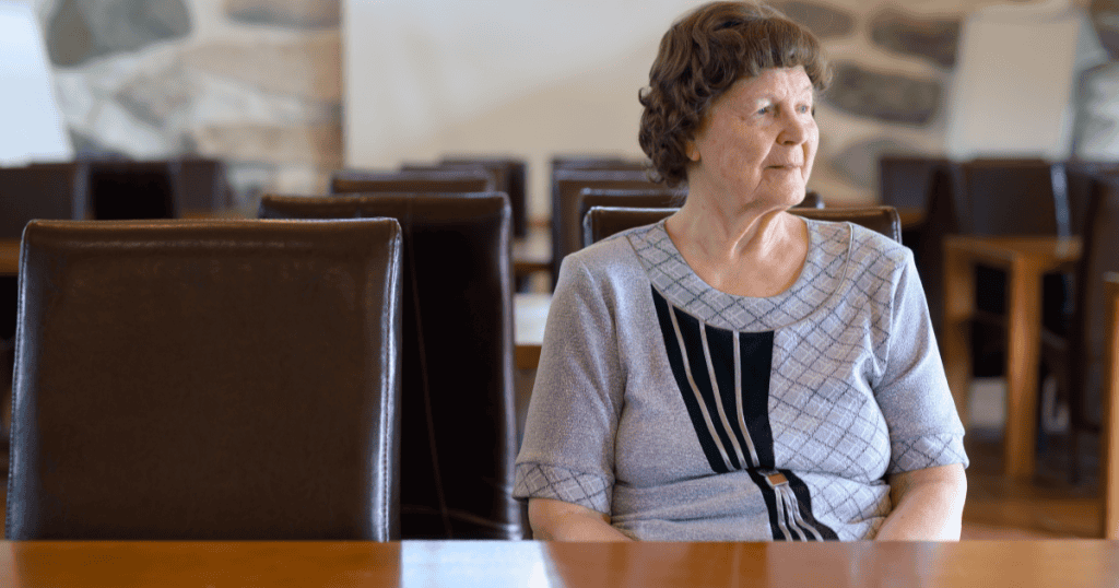 An older woman sits alone at a long table surrounded by empty chairs, gazing off to the side, reflecting how a narcissist's humiliation ultimately reveals the loneliness and fragile self-worth hidden beneath their image.