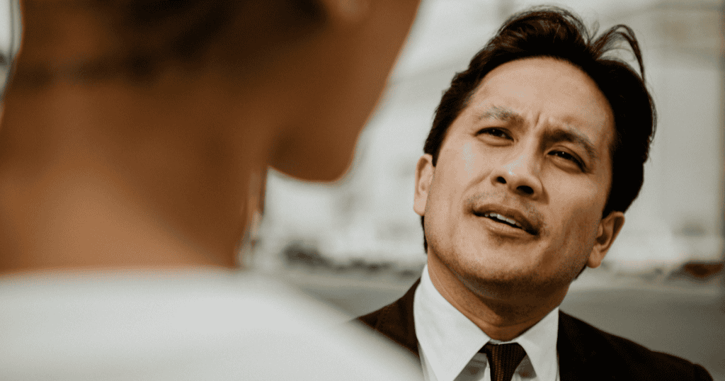 A man in a suit speaks to someone with a condescending, doubtful expression, illustrating the toxic phrases a narcissist relies on to slowly chip away at your confidence.