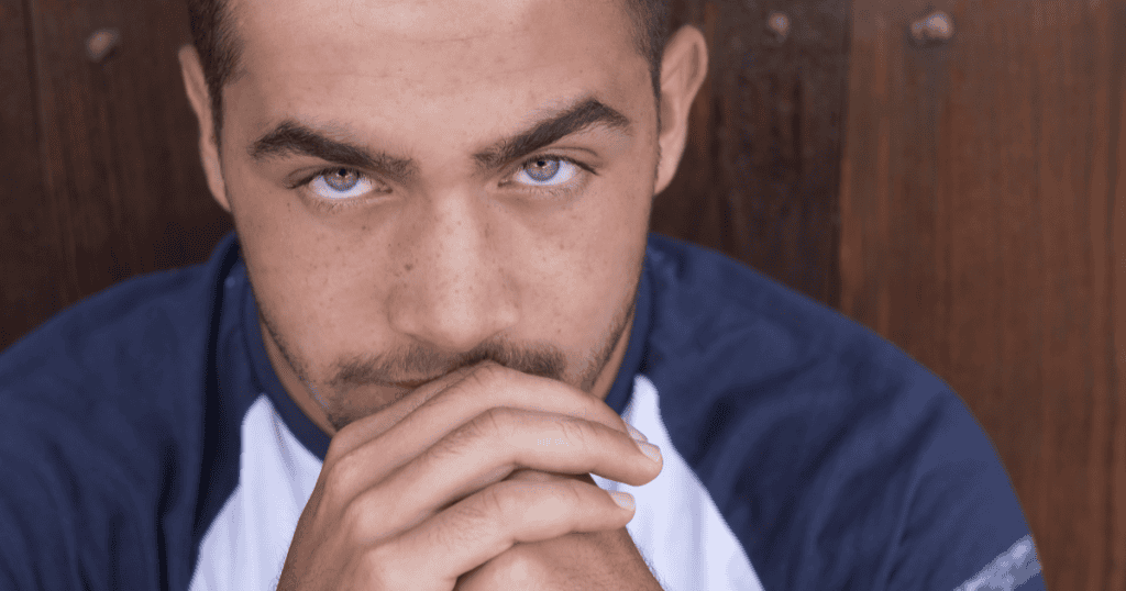 A young man stares intently from behind his clasped hands with a guarded, calculating look, hinting at the quiet, unusual habits that reveal a covert narcissist.