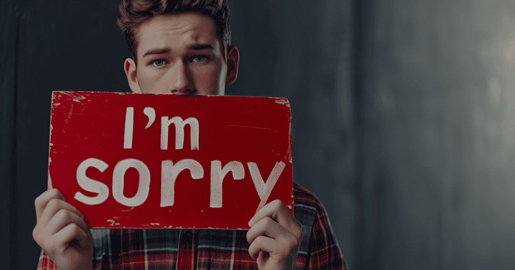 A young man hides behind a red “I’m sorry” sign with a hollow, sheepish look, illustrating how a covert narcissist’s apologies are a performance rather than genuine remorse.