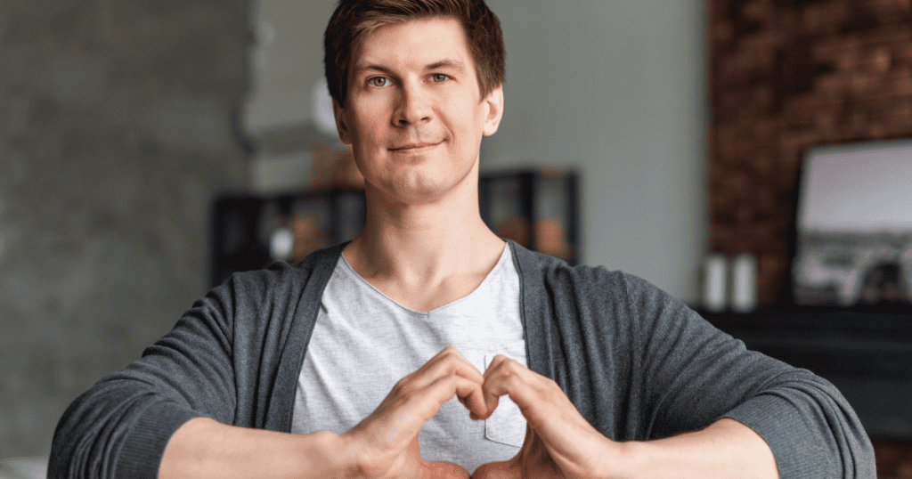 A man smiles gently while forming a heart with his hands, showing how a covert narcissist’s kind and caring first impression makes them so easy to miss at first.