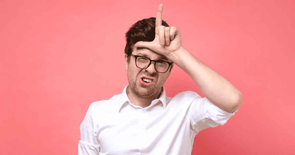 A man in glasses grimaces while making an “L” for loser on his forehead, illustrating how a covert narcissist takes any loss as a personal attack on their worth.