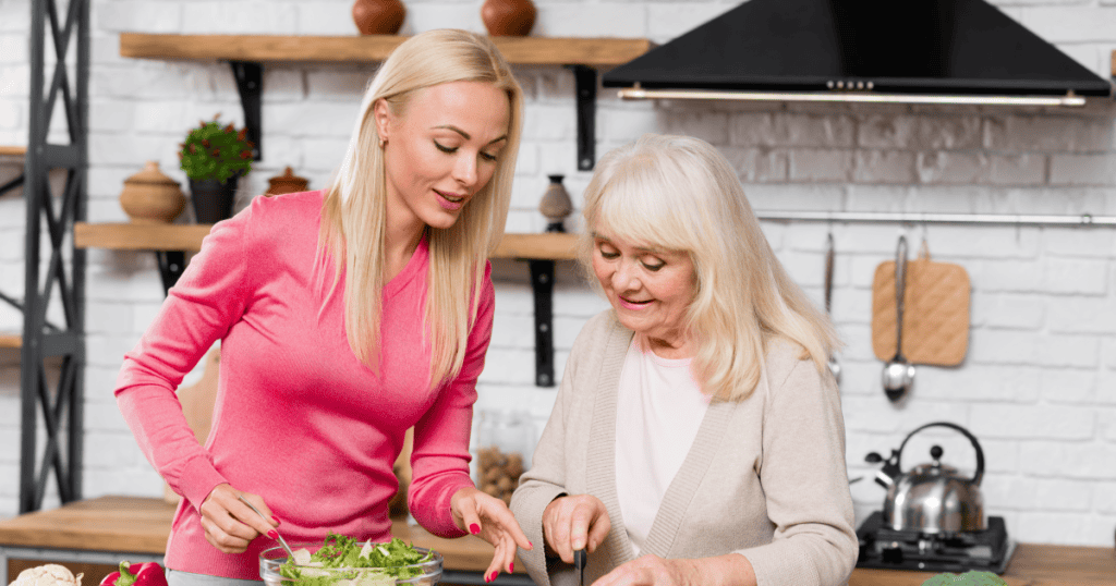 A younger woman leans over and points at what an older woman is chopping in the kitchen, illustrating how a covert narcissist turns small corrections into a constant habit of nitpicking.